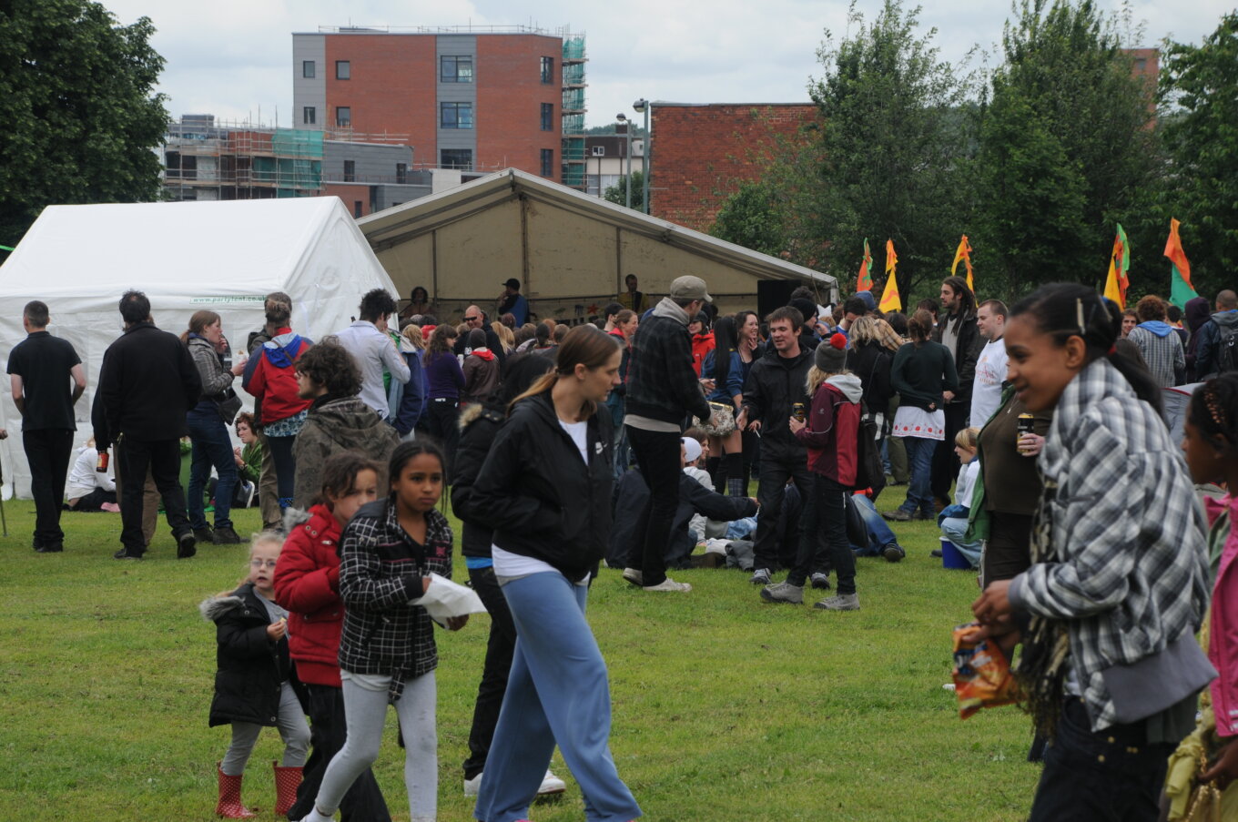 A group of people at an outdoor festival on grass, with tents in the background and buildings behind them.