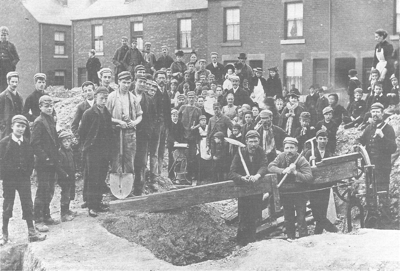 A black and white photograph of a group of mostly men and boys with tools and a hole in the ground.