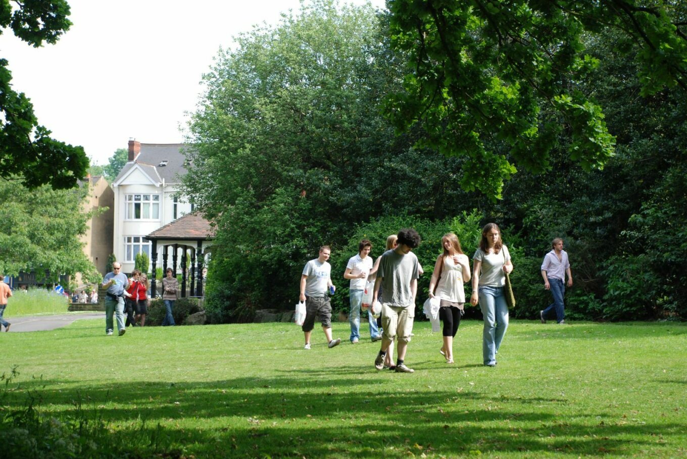 A group of young people walk through a green park, under trees, with a house in the background.