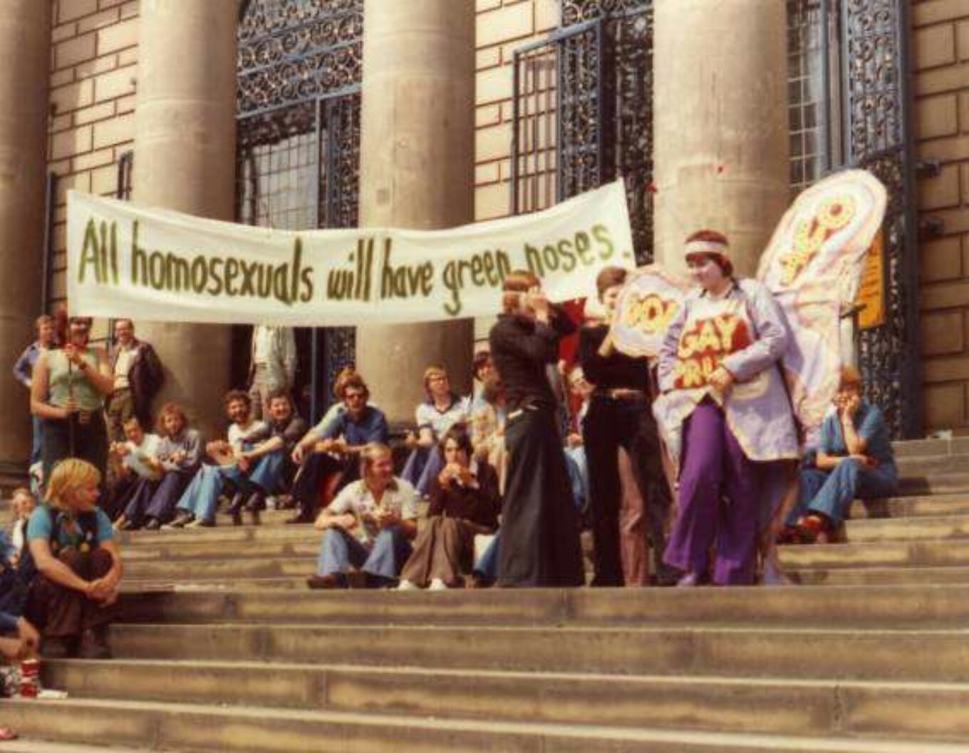 A group of people sitting and standing on the steps of City Hall One person is wearing fairy wings and a handmade t shirt that reads Gay Pride Some people hold a banner that reads All homosexuals will have green noses