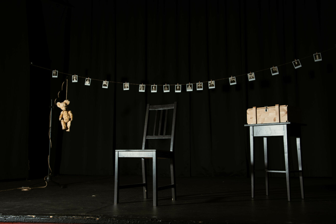 A dark stage with a teddy bear, an empty chair and a washing line with photographs