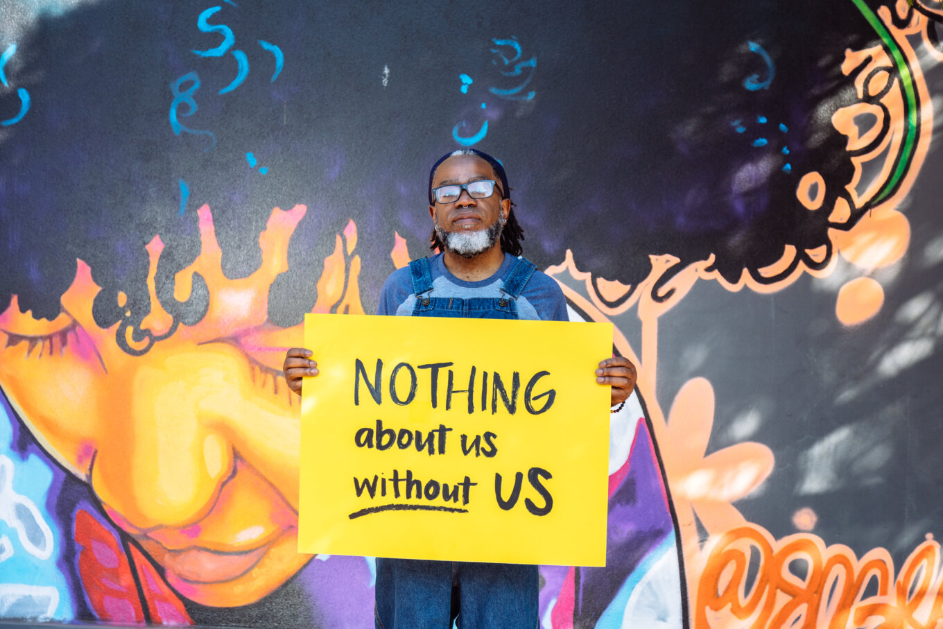 A Deaf Black man wearing glasses looks neutrally at the camera while holding a hand lettered sign declaring “NOTHING about us without US”.