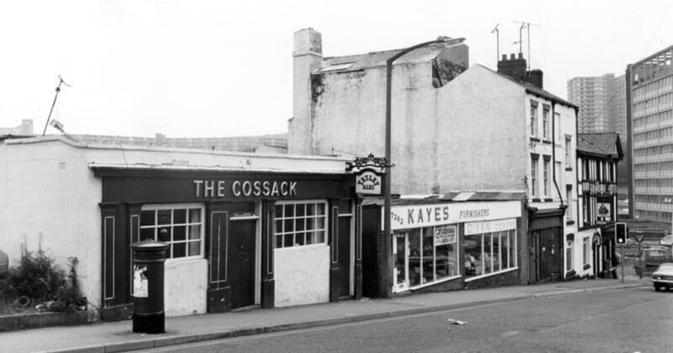 Side view from the road of a pub called The Cossack with a dark facade metallic lettering and a Tetley Ales hanging sign A furniture shop and some brutalist buildings are visible to the right of it