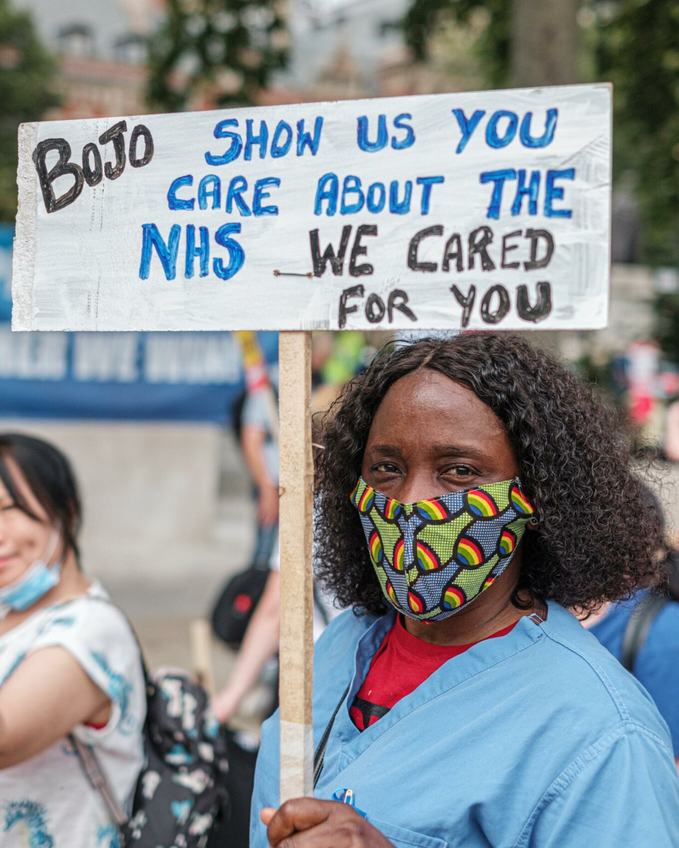 A Black woman wearing scrubs has a sign that says "BoJo show us you care about the NHS... we cared for you"