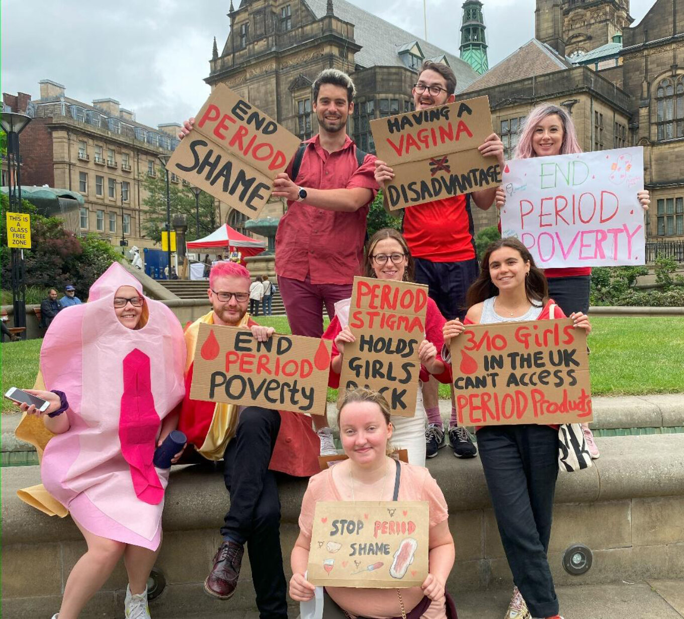 A group of young people in the Peace Gardens in Sheffield hold up handmade signs saying things like End Period Poverty and Period Stigma Holds Girls Back.