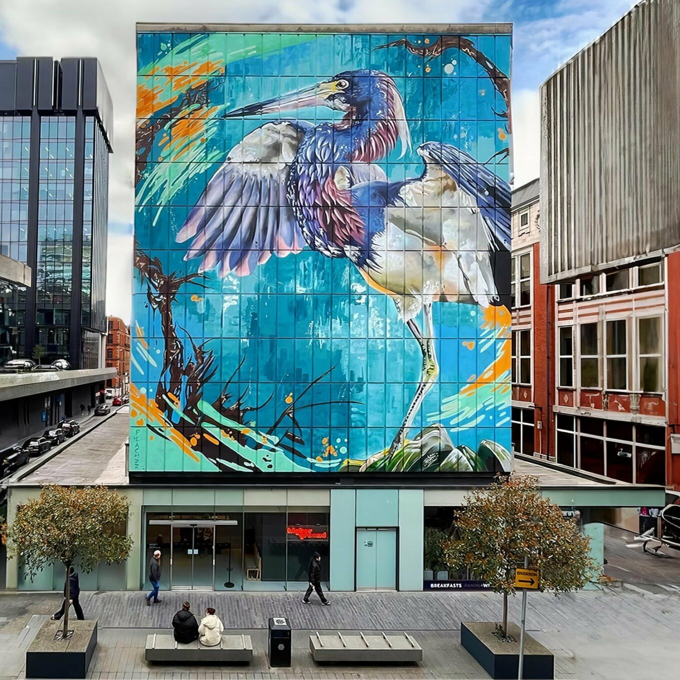 On a large city wall, there's a painting of a heron against a background of blues and greens.