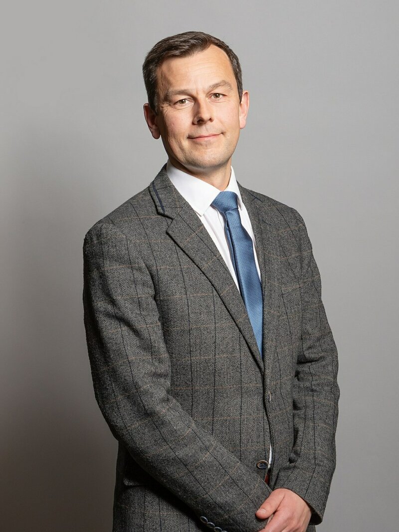 A white man wearing a grey suit in front of a grey wall