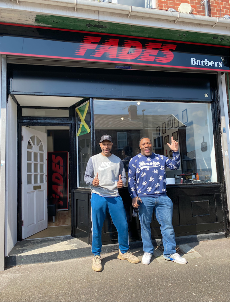 Two men stand in front of a shopfront painted in blue, with the word FADES in red.
