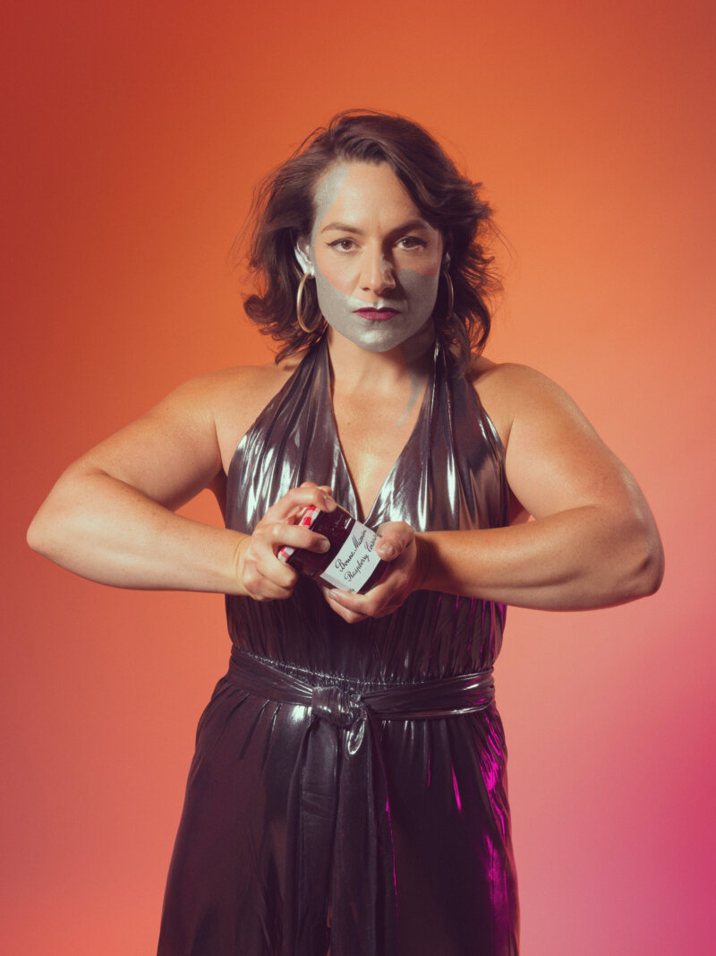 A woman with muscular arm stares at the camera while opening a jar of jam. She has metallic paint across her face.