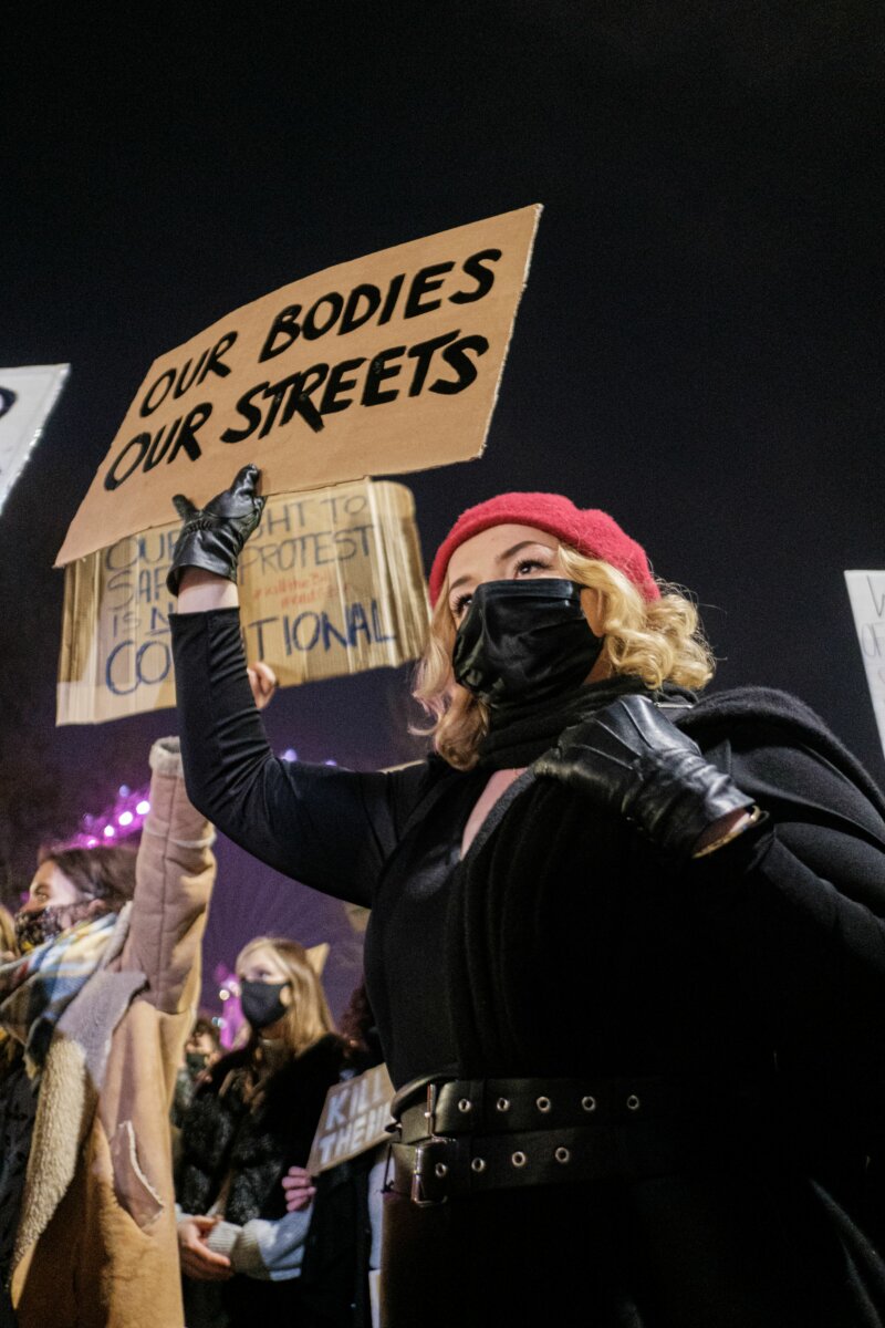 A woman with her face covered holds a hand-painted sign that says Our bodies our streets.